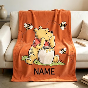May include: A soft orange fleece blanket featuring a yellow bear wearing a red shirt, sitting beside a honey pot. Three small bees with black and yellow stripes fly around the bear. The word NAME is printed in black capital letters at the bottom.