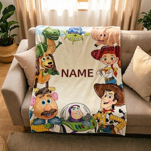 May include: A soft fleece blanket featuring a colourful print of various animated characters including a green dinosaur, a brown dog, a potato-shaped figure, a space ranger, a pig, a cowgirl, and a cowboy, with the word NAME printed in the centre.