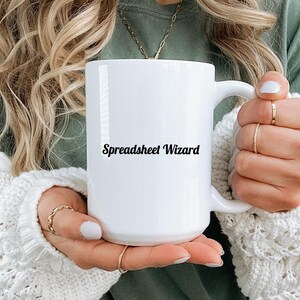 May include: A white ceramic mug featuring the black text Spreadsheet Wizard printed on the front. The mug has a standard cylindrical shape with a curved handle on the right side, providing a clear view of the printed lettering against the white surface.