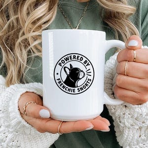以下が含まれることがあります： 中央にフレンチブルドッグのシルエットと「POWERED BY FRENCHIE SNORTS」という文字が黒い円形のグラフィックでプリントされた白いセラミック製マグカップ。マグカップは滑らかな質感で、右側に湾曲したハンドルが付いています。