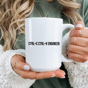 May include: A white ceramic mug featuring the black printed text CTRL+C CTRL+V ENGINEER. The mug has a smooth, cylindrical body with a curved handle on the right side, designed for holding hot beverages in a standard, functional style.