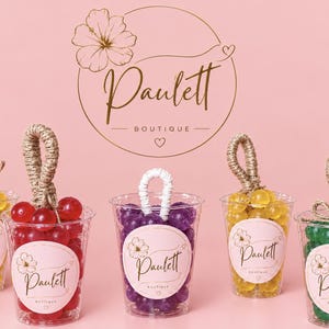 May include: Five clear plastic cups filled with colorful round beads in yellow, red, purple, and green. Each cup features a round sticker with the text Paulett Boutique and a floral icon. Four cups have jute rope handles, while one has a white handle.