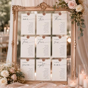 May include: A large ornate gold-toned rectangular frame displaying nine white table seating cards arranged in three rows. Each card features black text for table numbers one through nine, guest names, and a small circular brown wax seal at the top.