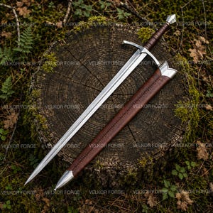 Può includere: Una spada in acciaio con lama a doppio taglio, guardia a croce e impugnatura rivestita in pelle marrone, accompagnata da un fodero in pelle marrone abbinato con puntale e bocchetta in metallo color argento, con il testo VELKOR FORGE visibile.