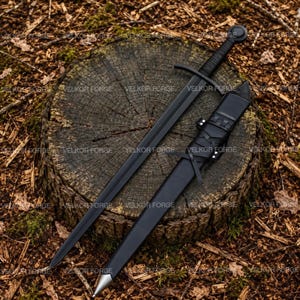 Puede incluir: Una espada negra mate con una guarda recta y un pomo circular, acompañada de una vaina negra a juego con detalles de correas de cuero y una punta de metal plateado. La espada está colocada en diagonal sobre un tocón de madera.