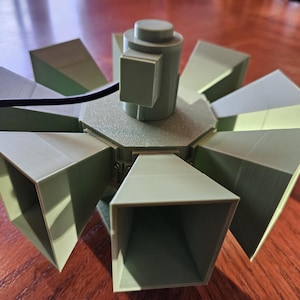 May include: A light green, 3D-printed mechanical device featuring a central cylindrical motor housing surrounded by eight radiating, funnel-shaped horn components. A thin black electrical wire extends from the top of the central motor unit.
