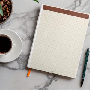Puede incluir: Un cuaderno con una página de puntos de color crema que presenta una barra de encabezado marrón con la etiqueta Notes. El cuaderno tiene un lomo blanco y un pequeño marcapáginas de cinta naranja. Un bolígrafo verde oscuro está al lado.