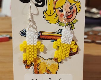 Op de afbeelding: Een paar banaanvormige oorbellen gemaakt van gele en witte plastic strijkkralen. De oorbellen hangen aan zilverkleurige metalen haakjes en worden gepresenteerd op een kaart met de letter Gg en een cartoonillustratie met de tekst EBearringsEtc.