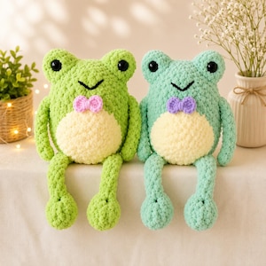 Op de afbeelding: Twee gehaakte knuffelkikkers met grote zwarte ogen en crèmekleurige buiken. De linker kikker is felgroen met een roze strikje, de rechter is mintgroen met een paars strikje. Beide hebben lange, bungelende armen en benen.