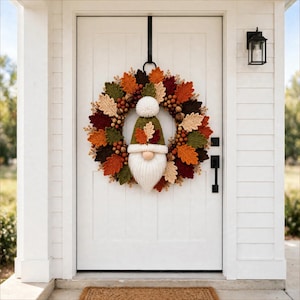 May include: A circular wreath featuring crocheted leaves in shades of orange, red, green, and beige, accented with small brown acorns. A central gnome figure wears a green pointed hat with a white pom-pom and a long, white, textured beard.