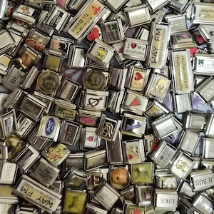 Könnte beinhalten: Eine Sammlung zahlreicher rechteckiger, modularer Edelstahl-Gliederarmband-Elemente, die jeweils mit verschiedenen bunten Emaille-Designs, Texten und Symbolen versehen sind und auf einem weißen Tablett mit Schmetterlings- und Sternenmuster liegen.