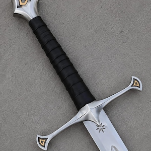 May include: A silver-toned metal sword featuring a black leather-wrapped grip, a crossguard with gold-colored triangular accents, and a matching pommel. The blade displays an engraved eight-pointed star near the hilt and a central fuller groove.