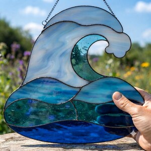 May include: A stained glass suncatcher shaped like a crashing ocean wave, featuring various shades of blue and white glass pieces joined by dark metal solder lines. The design includes a hanging chain at the top for display in a window.