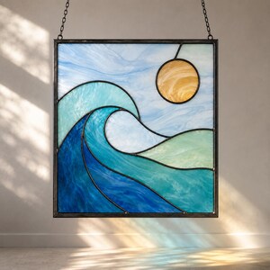 Puede incluir: Un panel de vidriera cuadrado que presenta una ola oceánica estilizada en tonos azul oscuro, turquesa y verde claro. Un sol circular de color ámbar se sitúa en la esquina superior derecha sobre un cielo azul pálido. Enmarcado en metal oscuro.