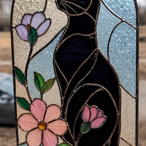 Op de afbeelding: Een boogvormig glas-in-loodpaneel met een zwarte kat in profiel. De kat is omringd door roze en paarse bloemen met groene bladeren. De achtergrond bestaat uit gestructureerde crèmekleurige en lichtblauwe glasstukken in een metalen omlijsting.