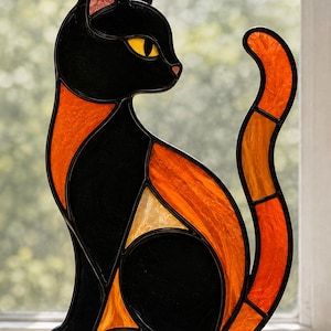 Puede incluir: Una figura de gato de vitral construida con paneles de vidrio negro, naranja y amarillo. El gato está representado en una posición sentada de perfil con una cola larga y curva. Las piezas de vidrio están unidas por líneas de metal oscuro.