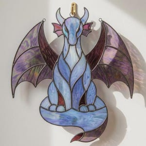 Op de afbeelding: Een glas-in-lood draak suncatcher met iriserende blauwe, paarse en violette glazen panelen die bijeengehouden worden door donkere metalen soldeerlijnen. De draak is zittend afgebeeld met gespreide vleugels en een gekrulde staart aan de basis.