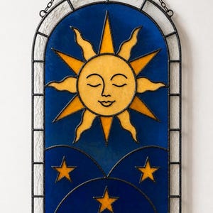 May include: An arched stained glass suncatcher featuring a yellow sun with a serene face at the top, surrounded by alternating straight and wavy rays. Below the sun, three yellow stars are set against a deep blue background with dark metal cames.