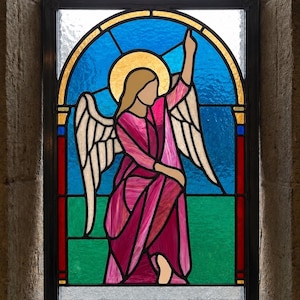 May include: A rectangular stained glass window panel featuring an angel with light-colored hair and a golden halo. The angel wears a pink robe and has white wings. The background consists of a blue sky above a green field, framed by a black border.