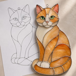 Könnte beinhalten: Ein Buntglas-Sonnenfänger in Form einer sitzenden Katze mit orangefarbenen, cremefarbenen und weißen Glasteilen, die durch silberne Metalllötlinien verbunden sind. Die Katze hat leuchtend grüne Augen und einen kleinen Metallring zum Aufhängen.