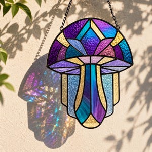 May include: A stained glass suncatcher shaped like a mushroom, featuring iridescent purple, blue, and yellow glass panels held together by dark metal soldering. The piece hangs from a black metal chain and casts colorful light reflections.