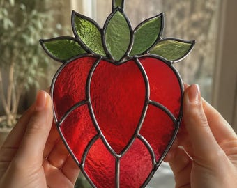 May include: A stained glass suncatcher shaped like a strawberry, featuring vibrant red glass segments for the fruit body and textured green glass pieces for the leaves at the top, all held together by dark metal solder lines in a decorative pattern.