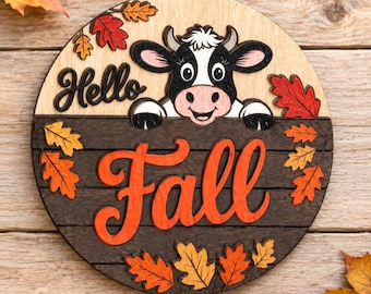 May include: A round wooden sign featuring a black and white cow illustration with the text Hello Fall. The top half is light wood, the bottom half is dark brown horizontal planks, and the design is decorated with orange and red autumn leaves.