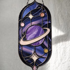 Könnte beinhalten: Ein ovaler Sonnenfänger aus Buntglas mit einem zentralen violetten Planeten mit blassrosa Ring, umgeben von kleineren Himmelskörpern in Rosa, Orange und Gelb, vor einem Hintergrund aus wirbelndem tiefem Blau und Violett mit Sternakzenten.