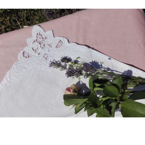 May include: A white linen handkerchief featuring an intricate cutwork embroidery pattern along the corner edge. A sprig of purple lavender and a single pink rosebud with green leaves are placed across the fabric surface.