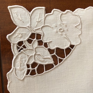 Può includere: Un tovagliolo di lino color bianco sporco caratterizzato da un intricato disegno angolare di fiori e foglie ricamati con dettagli a giorno. I bordi del tessuto sono smerlati e il ricamo è eseguito con un filo bianco sporco abbinato.