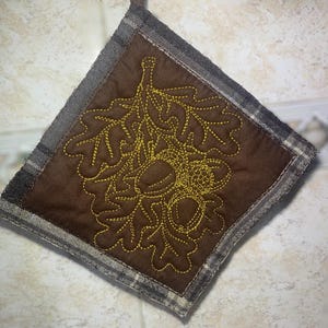 May include: A square potholder featuring a brown center panel with a yellow embroidered design of oak leaves and acorns. The border consists of a gray and white plaid fabric. The item is oriented in a diamond shape with a hanging loop at the top.