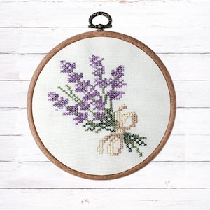 May include: A circular wooden embroidery hoop containing white fabric with a cross-stitch design of a lavender bouquet. The lavender flowers are rendered in shades of purple, with green stems and leaves, tied together with a tan-colored ribbon.