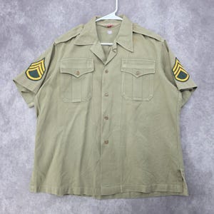 May include: A short-sleeved khaki military-style shirt featuring a pointed collar, a button-down front, and two chest pockets with buttoned flaps. Each sleeve displays a green and yellow chevron patch, and the shoulders include epaulets.