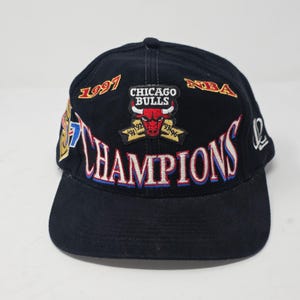 Peut inclure: Une casquette de baseball noire avec le logo des Chicago Bulls, le texte 1997, NBA et CHAMPIONS brodé en rouge, blanc et or. La casquette affiche les années de championnat 91, 92, 93, 94, 95, 96 sur l'emblème avant et un logo sur le côté.