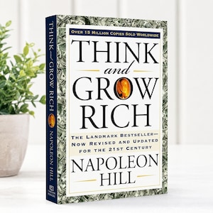 Puede incluir: Un libro de tapa dura titulado Think and Grow Rich de Napoleon Hill. La portada tiene un fondo blanco con texto negro, un gráfico de llama central y un borde con billetes de dólar. El lomo es de color azul oscuro con texto blanco.
