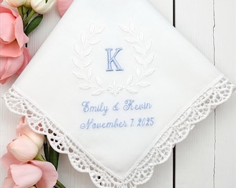 Può includere: Un fazzoletto quadrato bianco con un bordo in pizzo smerlato e ricami azzurri. Il centro mostra una lettera K all'interno di una corona d'alloro, con il testo Emily & Kevin e la data November 7, 2025, ricamati sotto in blu.
