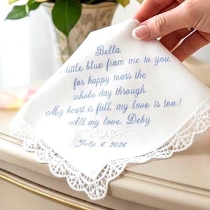 May include: A white fabric handkerchief with a scalloped lace border, featuring blue embroidered text that reads: Bella, little blue from me to you, for happy tears the whole day through. My heart is full, my love is too! All my love, Deby, July 6, 2026.