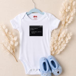 May include: A white short-sleeved baby bodysuit featuring a black graphic of a computer code window with the text #include <stdio.h> int main() { printf("Hello, world!"); return 0; }. The bodysuit is displayed on a light blue hanger with light blue booties.