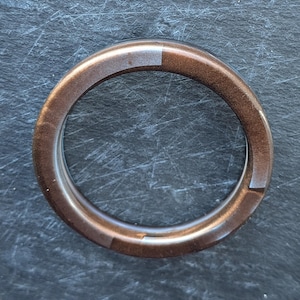 Può includere: Un anello circolare caratterizzato da una finitura liscia e lucida con una tonalità metallica marrone ramato. La fascia presenta sottili variazioni nell'intensità del colore, passando da sfumature più chiare a più scure di bronzo.