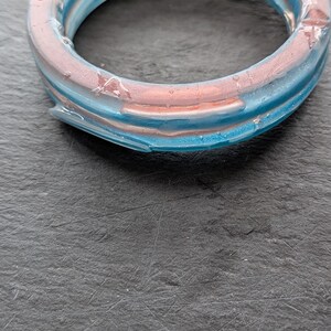 May include: A circular bangle bracelet crafted from translucent resin, featuring a layered design with a core of metallic copper-toned material encased within a clear, bright blue outer shell that has a smooth, glossy, and reflective finish.