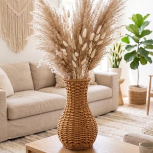 May include: A tall, woven rattan vase with a narrow neck and a rounded base, filled with a large arrangement of fluffy, beige pampas grass plumes and smaller, white, oval-shaped bunny tail dried flowers, creating a textured, natural botanical display.