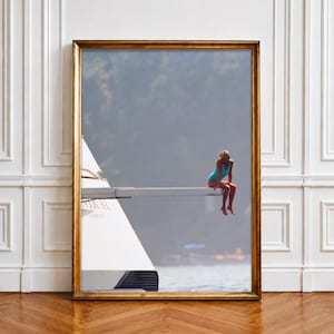 May include: A framed photographic print featuring a person in a bright blue one-piece swimsuit sitting on the edge of a white diving board attached to a large yacht. The yacht has a white hull with the word "KISMET" visible on the side.