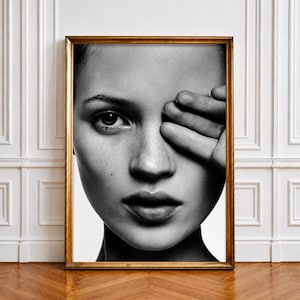 Könnte beinhalten: Ein schwarz-weiß Fotodruck in einem goldenen Rahmen zeigt ein Nahaufnahme-Porträt eines Gesichts, bei dem ein Auge sichtbar ist und das andere von einer Hand verdeckt wird. Das Bild zeigt detaillierte Hautstrukturen und einen hohen Kontrast.