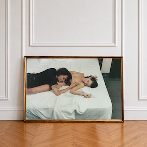 May include: A rectangular framed wall art print featuring a photographic scene of two individuals lying together on a white bed. The image shows one person in a black shirt embracing another person, captured in a horizontal orientation with a gold frame.