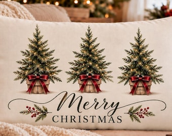 Puede incluir: Un cojín rectangular de color crema con tres árboles de hoja perenne en macetas, decorados con luces y lazos de color rojo oscuro. Debajo de los árboles, el texto Merry CHRISTMAS está impreso en letras negras, junto a pequeñas ramas con bayas rojas.