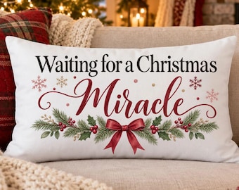 Puede incluir: Un cojín rectangular blanco con el texto negro Waiting for a Christmas, la palabra Miracle en letra cursiva roja y una ilustración decorativa de ramas de pino verdes, bayas rojas y un lazo rojo centrado debajo.