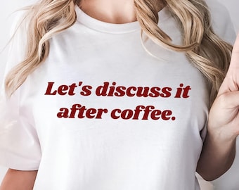 Puede incluir: Una camiseta blanca de manga corta con el texto Let's discuss it after coffee. impreso en una fuente serif de color rojo oscuro en el pecho. La camiseta tiene cuello redondo y un corte holgado, combinada con pantalones vaqueros azules.