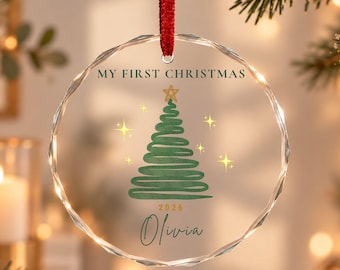 Puede incluir: Un adorno circular de cristal transparente con borde facetado, colgado de una cinta roja. Presenta un árbol de Navidad verde dibujado a mano con una estrella dorada, el texto "MY FIRST CHRISTMAS", el año "2026" y el nombre "Olivia" en negro.
