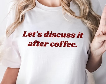 Puede incluir: Una camiseta blanca de manga corta con el texto Let's discuss it after coffee. impreso en una fuente serif de color rojo oscuro en el pecho. La camiseta tiene cuello redondo y un corte holgado, combinada con pantalones vaqueros azules.