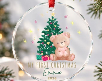 Puede incluir: Un adorno navideño de cristal circular y transparente que cuelga de una cinta roja. El diseño presenta un árbol de Navidad verde, un oso de peluche marrón claro con un lazo rosa y el texto "MY FIRST CHRISTMAS Emma 2026" en dorado y verde.